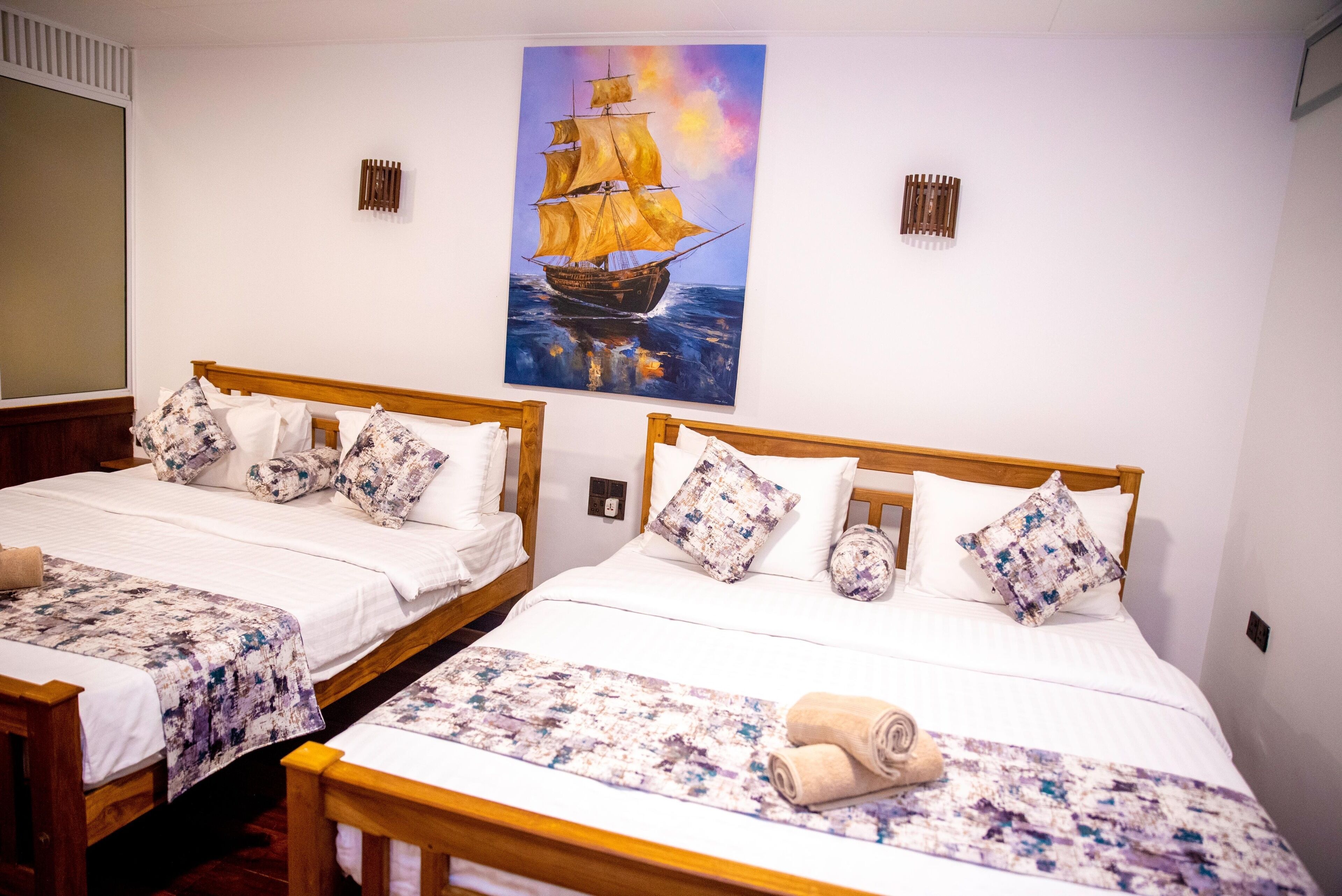 Une chambre d’hôtel avec deux lits en bois, du linge de lit blanc, des coussins à motifs, un tableau représentant un navire accroché au mur et une fenêtre avec des stores.