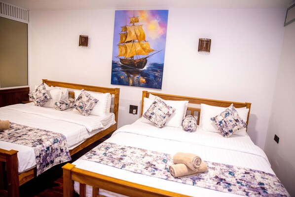 Ein Hotelzimmer mit zwei hölzernen Betten, weißer Bettwäsche, gemusterten Kissen, einem Gemälde eines Schiffes an der Wand und einem Fenster mit Jalousien.