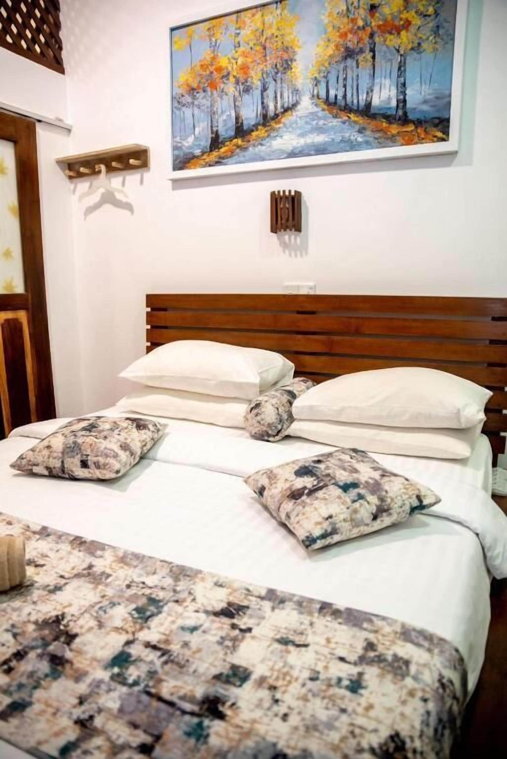 A neatly made bed with floral-patterned bedding and pillows, a wooden headboard, and a painting of a tree-lined path on the wall.
