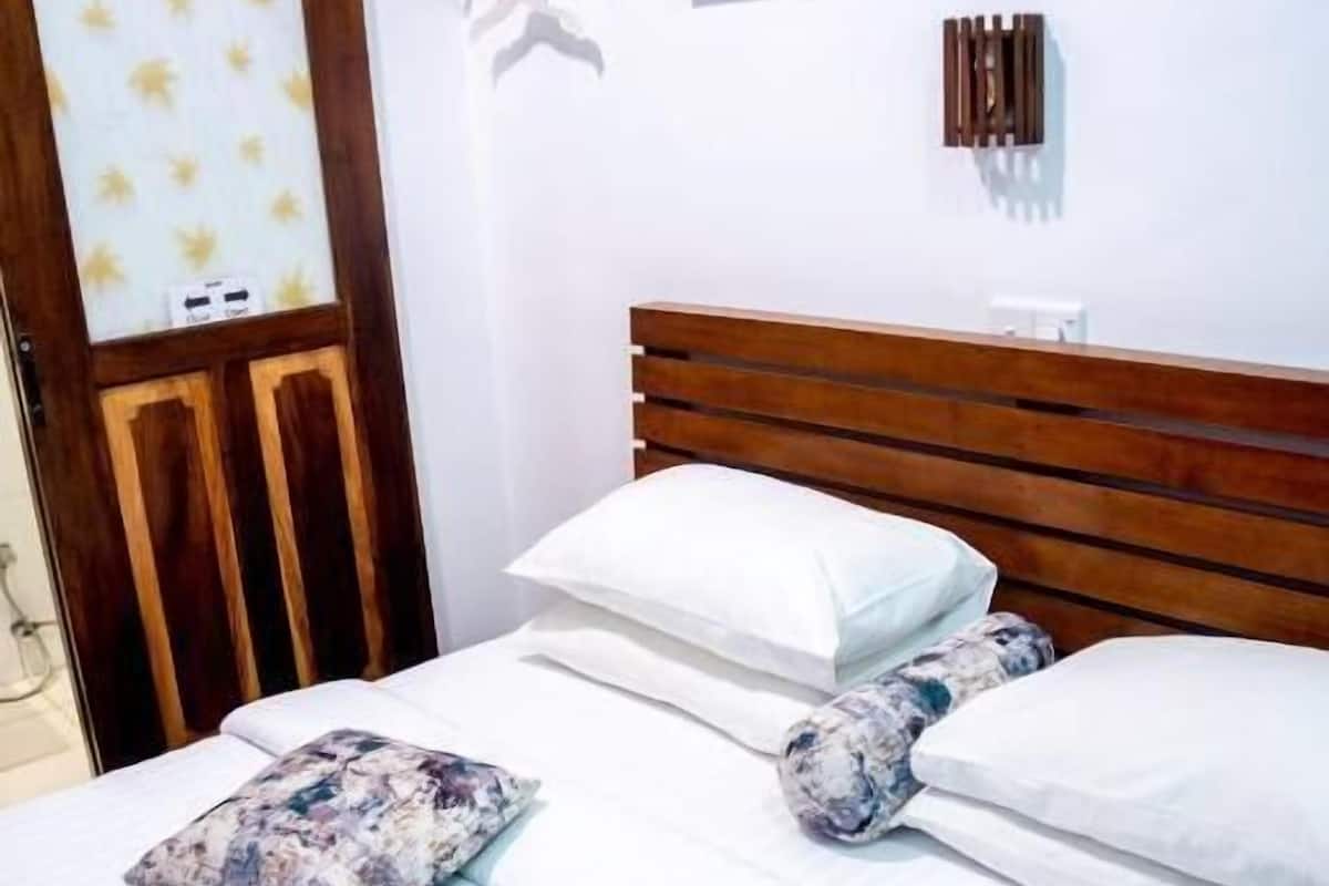 Una cama bien hecha con ropa blanca y almohadas estampadas, un cabecero de madera y un cuadro de un sendero forestal sobre ella.