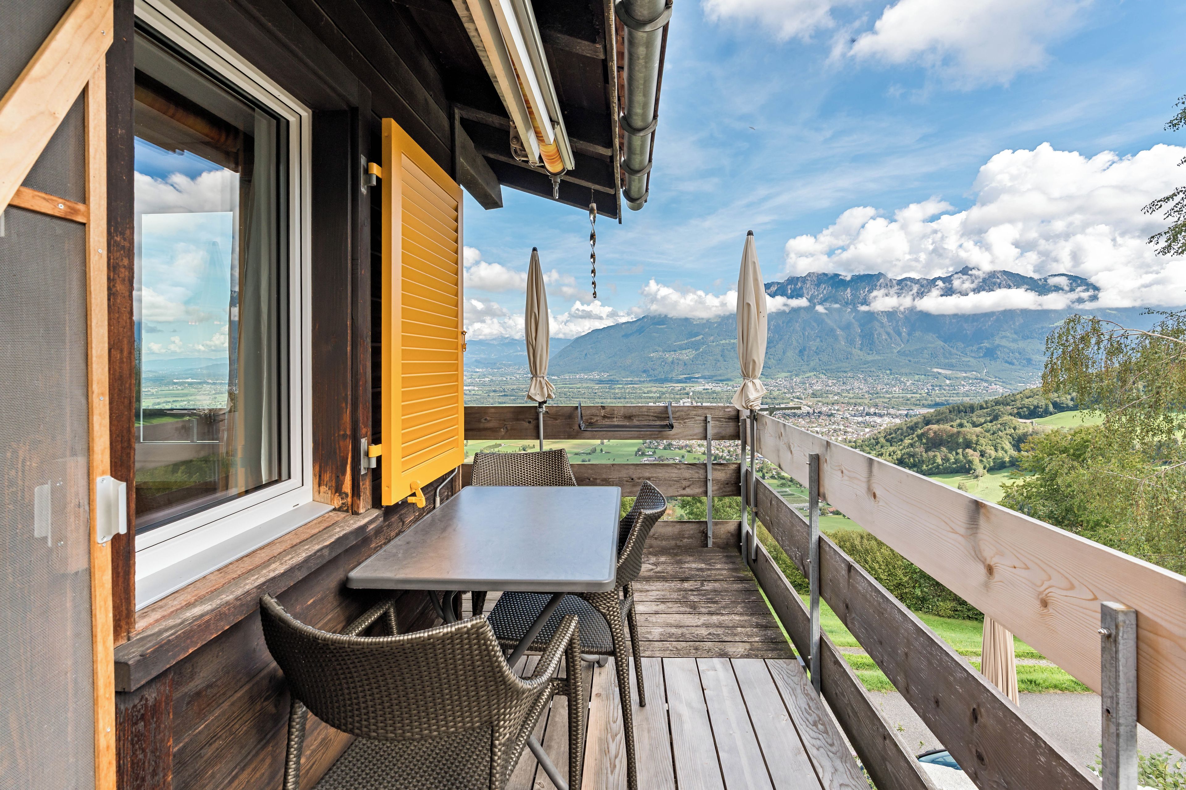 Een houten terras met tafel en stoelen, met uitzicht op een bergachtig landschap.