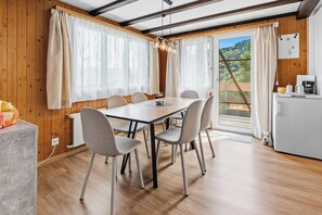 Ein Essbereich mit Tisch und Stühlen, Holzwänden, einem fenster mit Vorhängen und Blick auf einen bewaldeten Hügel.
