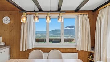 A dining area with a wooden interior, a large window with curtains, a clock on the wall, and a view of mountains.