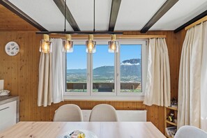 A dining area with a wooden interior, a large window with curtains, a clock on the wall, and a view of mountains.