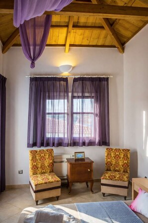 Ein Zimmer mit einem Bett, zwei Sesseln, einem kleinen Tisch mit einer Uhr und einem Fenster mit violetten Vorhängen.