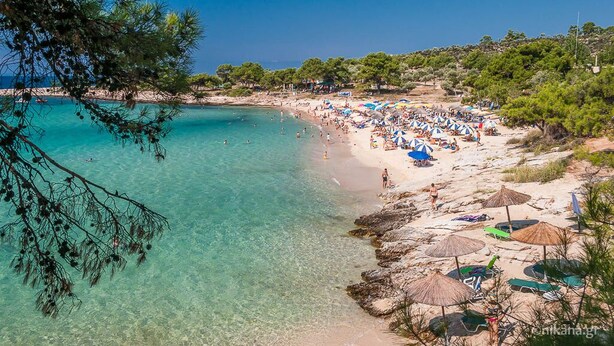 Une plage à l’eau turquoise et limpide, bordée de nombreux parasols, où les gens profitent du soleil.
