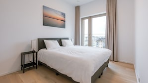 Ein modernes Schlafzimmer mit einem großen Bett, einem Nachttisch, einer Stehlampe und Blick auf die Stadt durch ein großes Fenster.