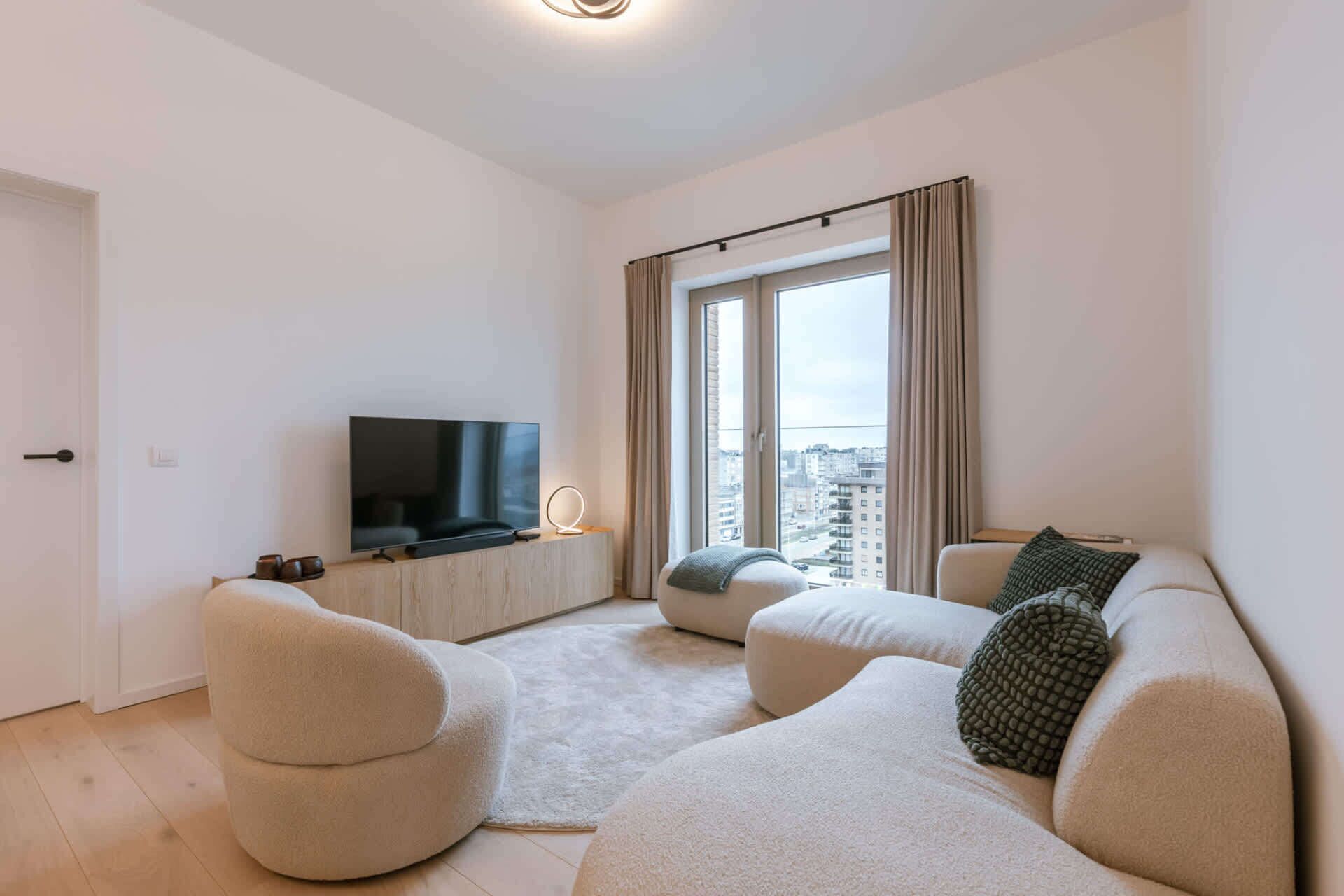 Een moderne woonkamer met een flatscreen televisie, twee banken, een salontafel en een groot raam met gordijnen.