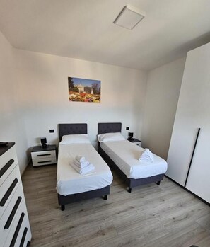 Una habitación de hotel moderna con dos camas individuales, una mesita de noche con lámpara, un armario y un cuadro en la pared.