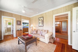 A living room with a sofa, coffee table, and a ceiling fan. There is a wooden floor and a door leading to another room. - Umetsu, Hanalei's Timeless Charm w/ AC TVNC#5096 (Hanalei)