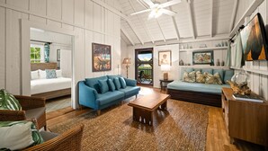 A spacious living room with a high ceiling, a ceiling fan, a blue sofa, a wooden coffee table, wicker chairs, and a view through a glass door to the outdoors. - Palaka Cottage, A Timeless Hanalei Stay TVNC#1091 (Hanalei)