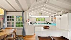 A kitchen with a white island, wooden chairs, a blue rug, and a view of greenery outside. - Palaka Cottage, A Timeless Hanalei Stay TVNC#1091 (Hanalei)