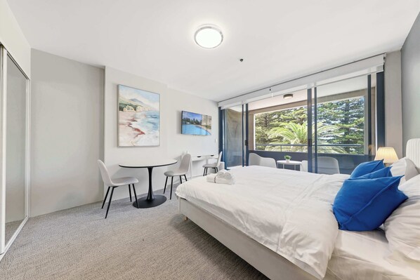 A modern hotel room with a bed, a desk, chairs, and a large window with a view of greenery.