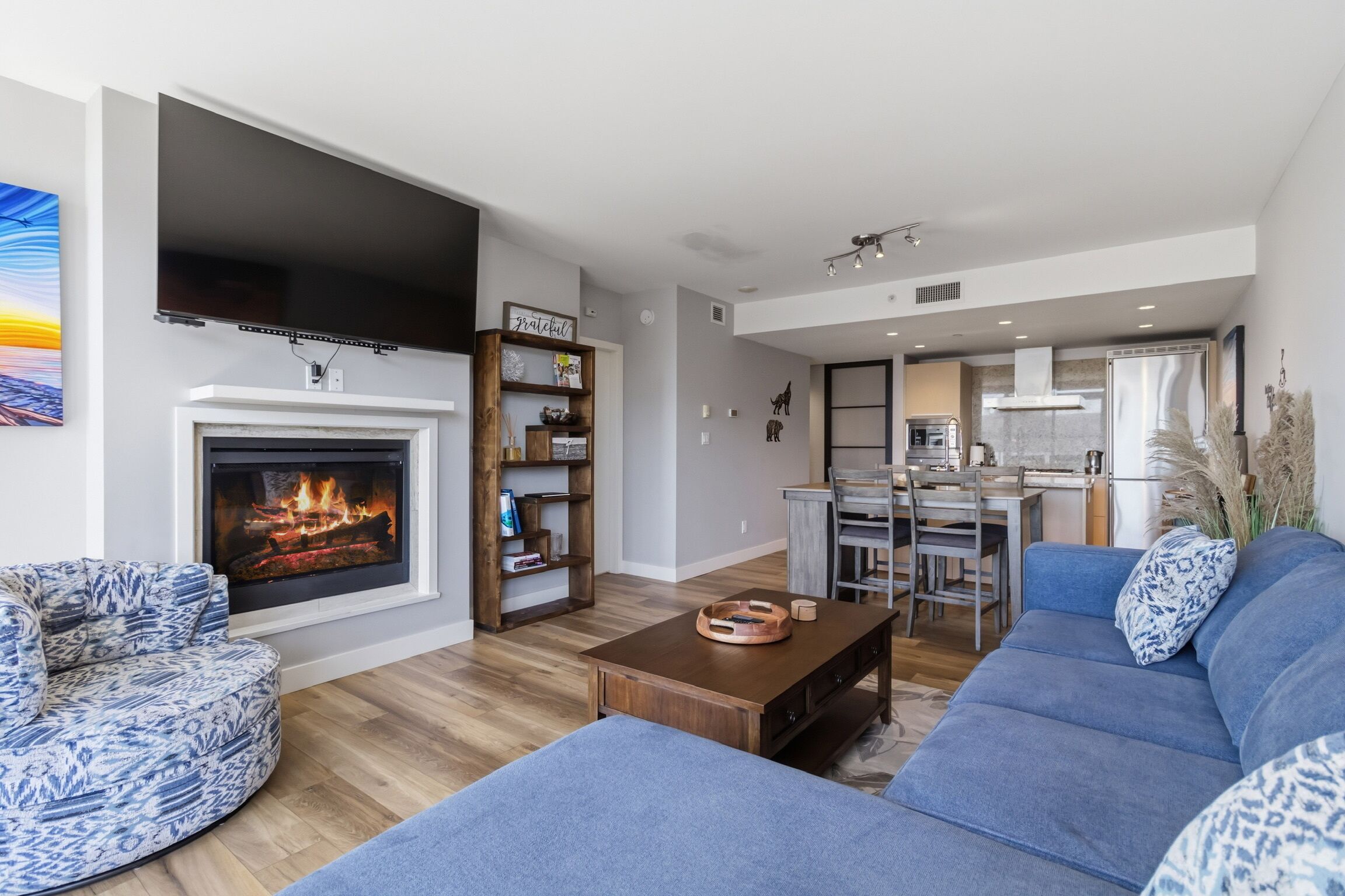 Een moderne woonkamer met een open haard, een blauw gestreept bankstel, een houten salontafel, en een keukenhoek met een eettafel en stoelen.