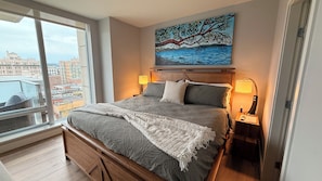 Un dormitorio con una cama grande, dos lámparas de noche, un cuadro sobre la cama y vistas a edificios a través de una ventana.