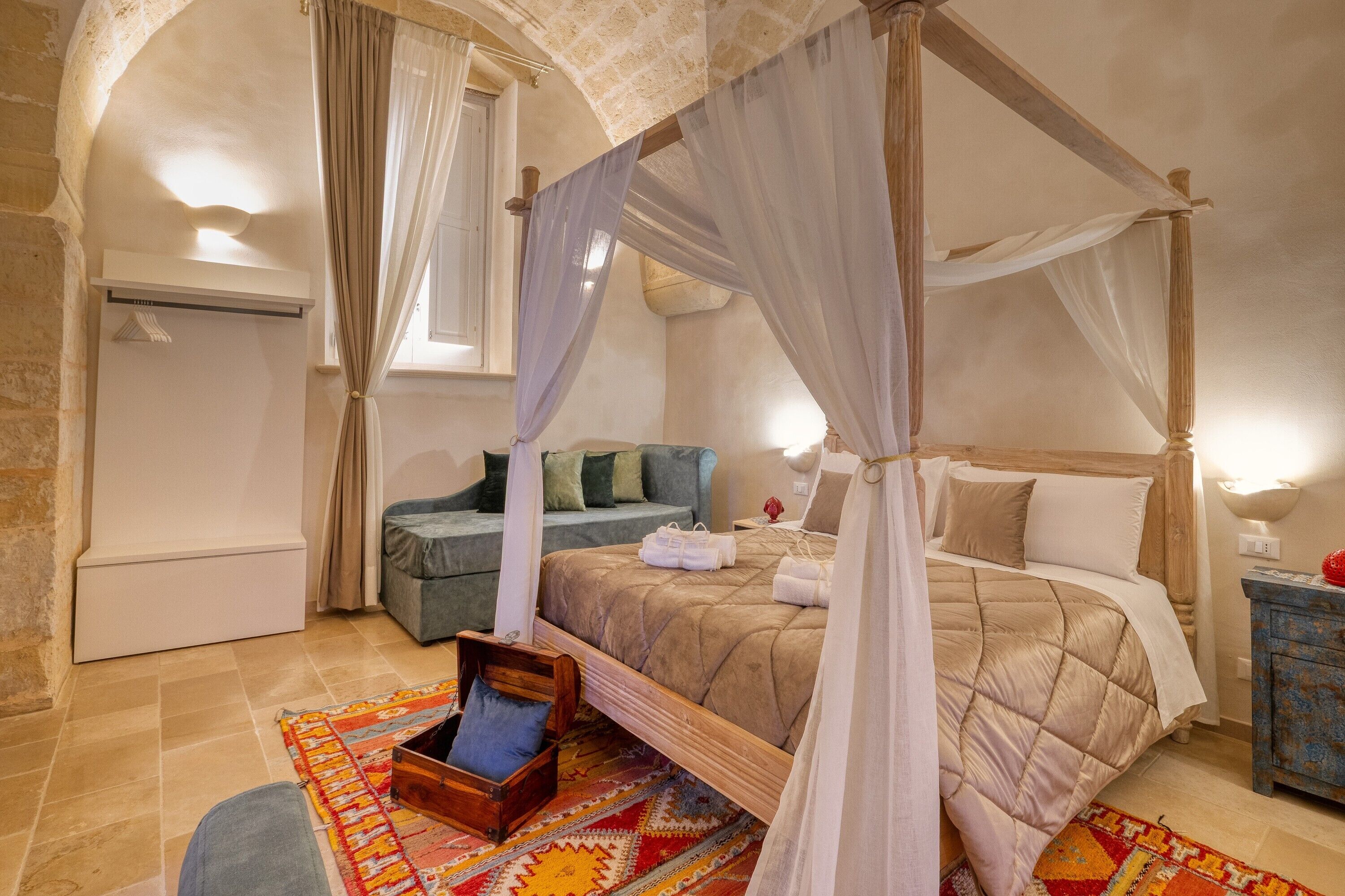 Ein Zimmer mit Stehwänden und einem Fenster, ausgestattet mit einem Himmelbett, einer Couch, einer kleinen Holzschatulle und einem gemusterten Teppich.