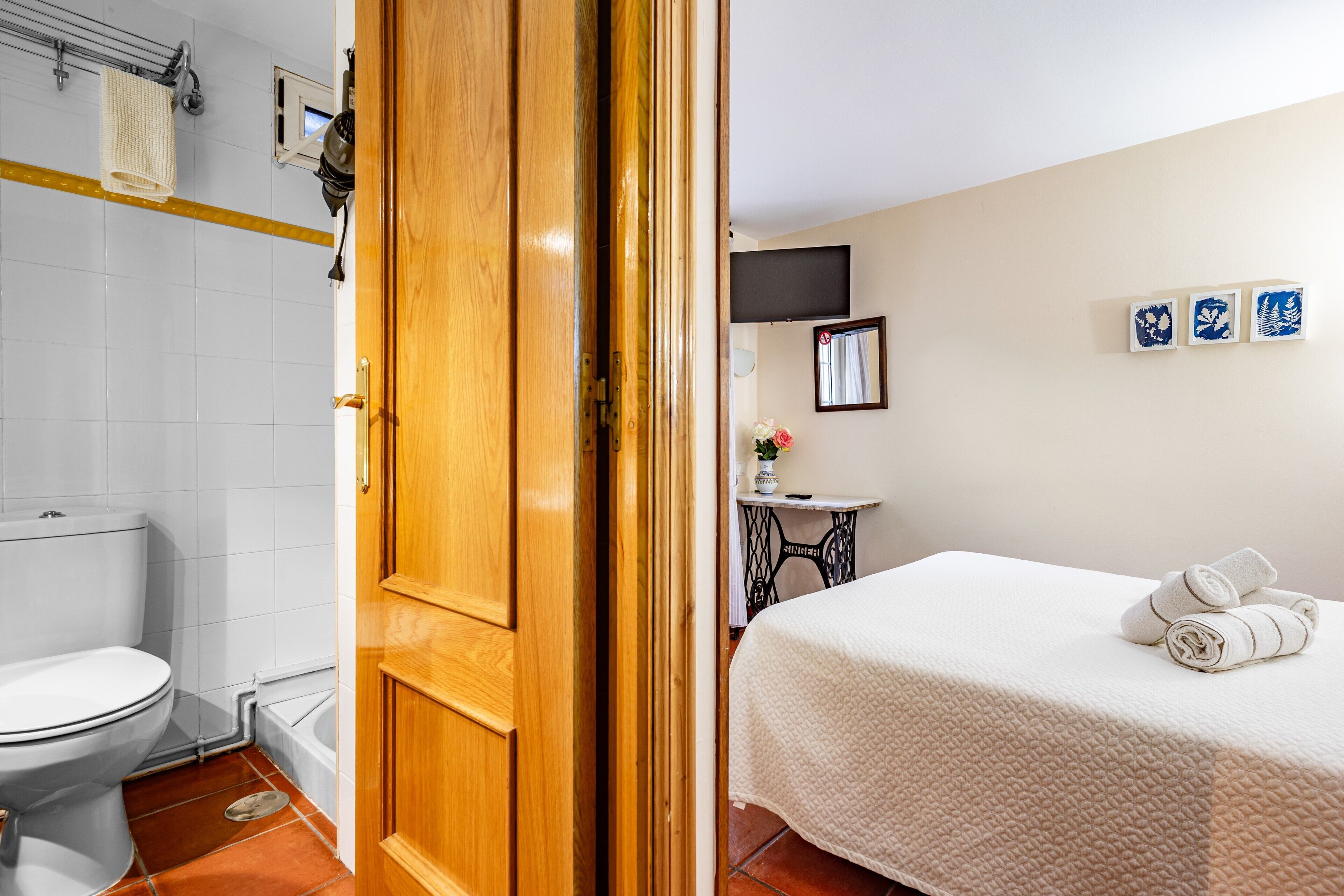Une chambre d’hôtel avec une porte en bois, un lit blanc avec des serviettes pliées, des toilettes blanches et une petite table sur laquelle se trouve un vase.