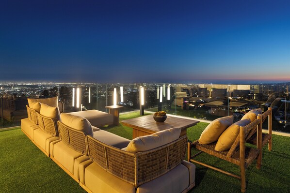 A rooftop terrace with a sofa set, a table, and a view of the cityscape at dusk.