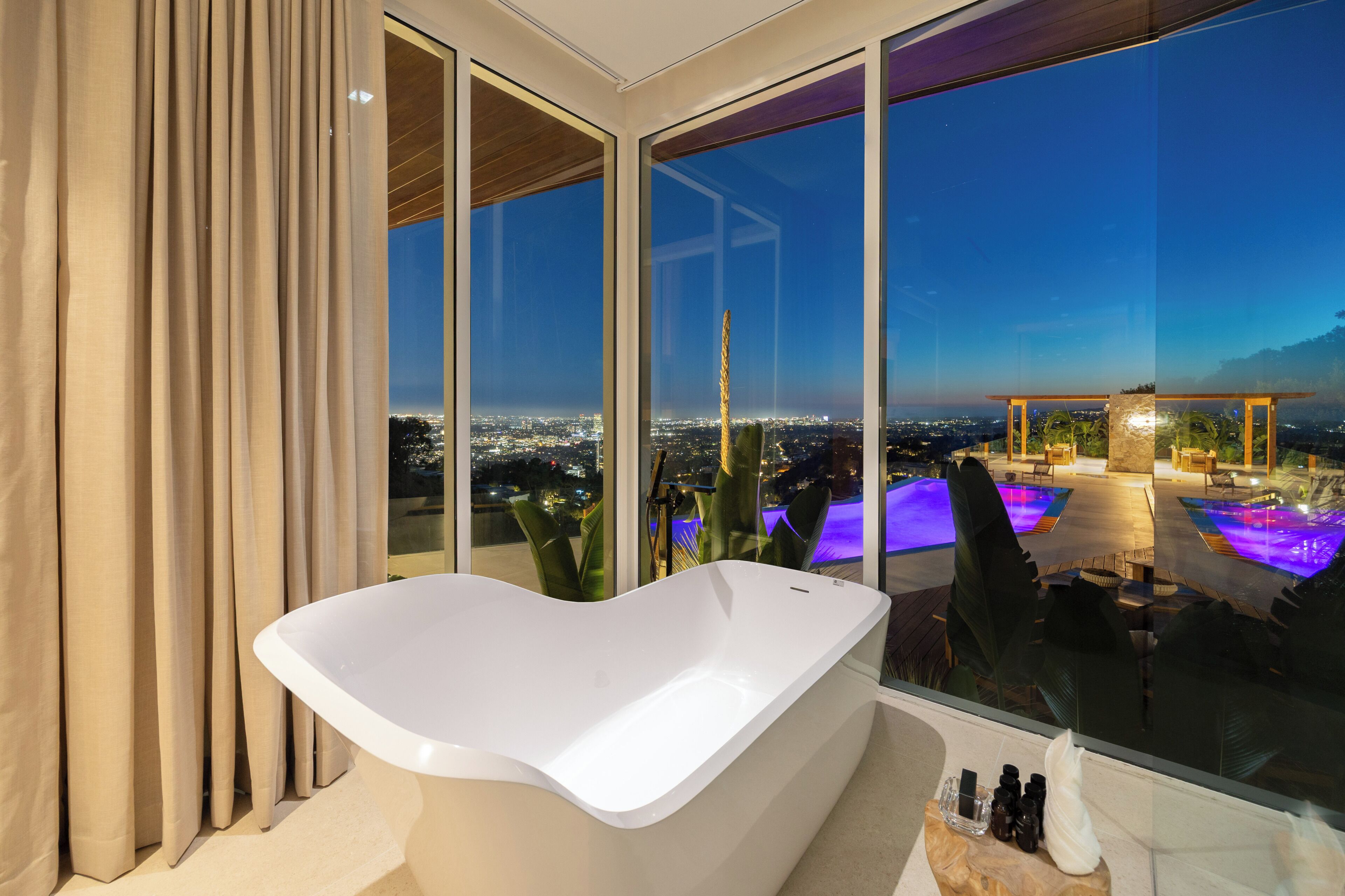 A modern bathroom with a freestanding bathtub, large windows, and a view of a pool and cityscape.