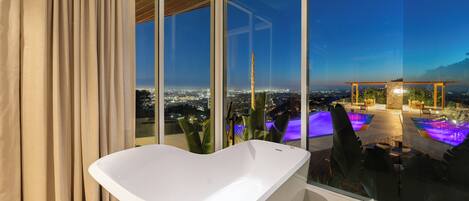 A modern bathroom with a freestanding bathtub, large windows, and a view of a pool and cityscape.