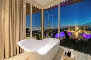 A modern bathroom with a freestanding bathtub, large windows, and a view of a pool and cityscape.