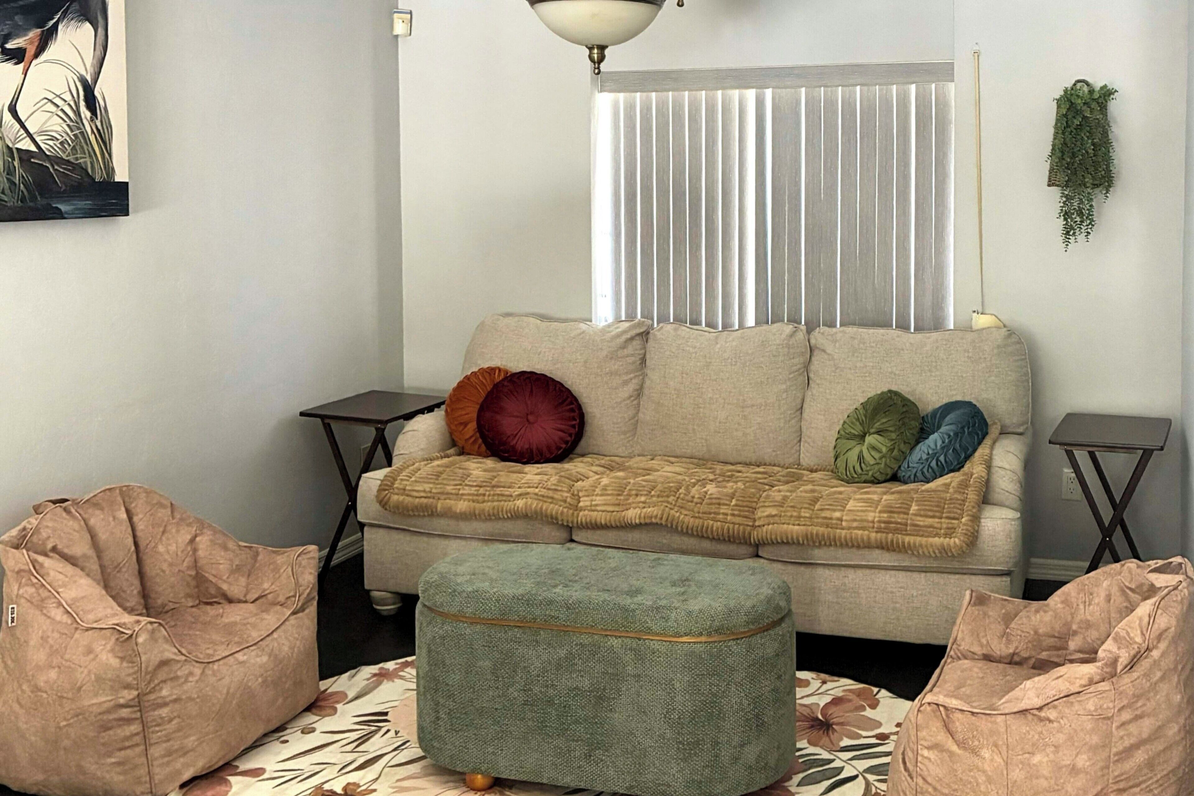 Un salon comprenant un canapé, un pouf, des fauteuils en forme de sac, un tableau, une fenêtre avec des stores, une plante suspendue et un lampadaire.