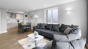 A modern living room with a grey sofa, a dining table with chairs, a kitchen in the background, and a large window. - The Isleworth 2BR 2BA with Gated Parking & Balcony (Isleworth)