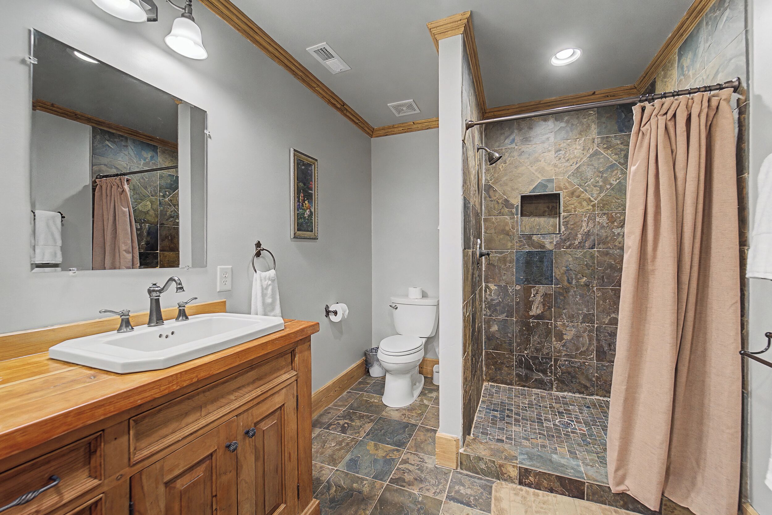 Une salle de bain avec un meuble-lavabo en bois, un lavabo blanc, des toilettes et un espace douche avec un mur en pierre.