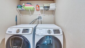 A laundry area with two Samsung washing machines, a shelf with folded towels and laundry detergent, and a hose connected to a power outlet. - 2 Mi to Ocean! Family-friendly Emerald Coast Haven (Fort Walton Beach)