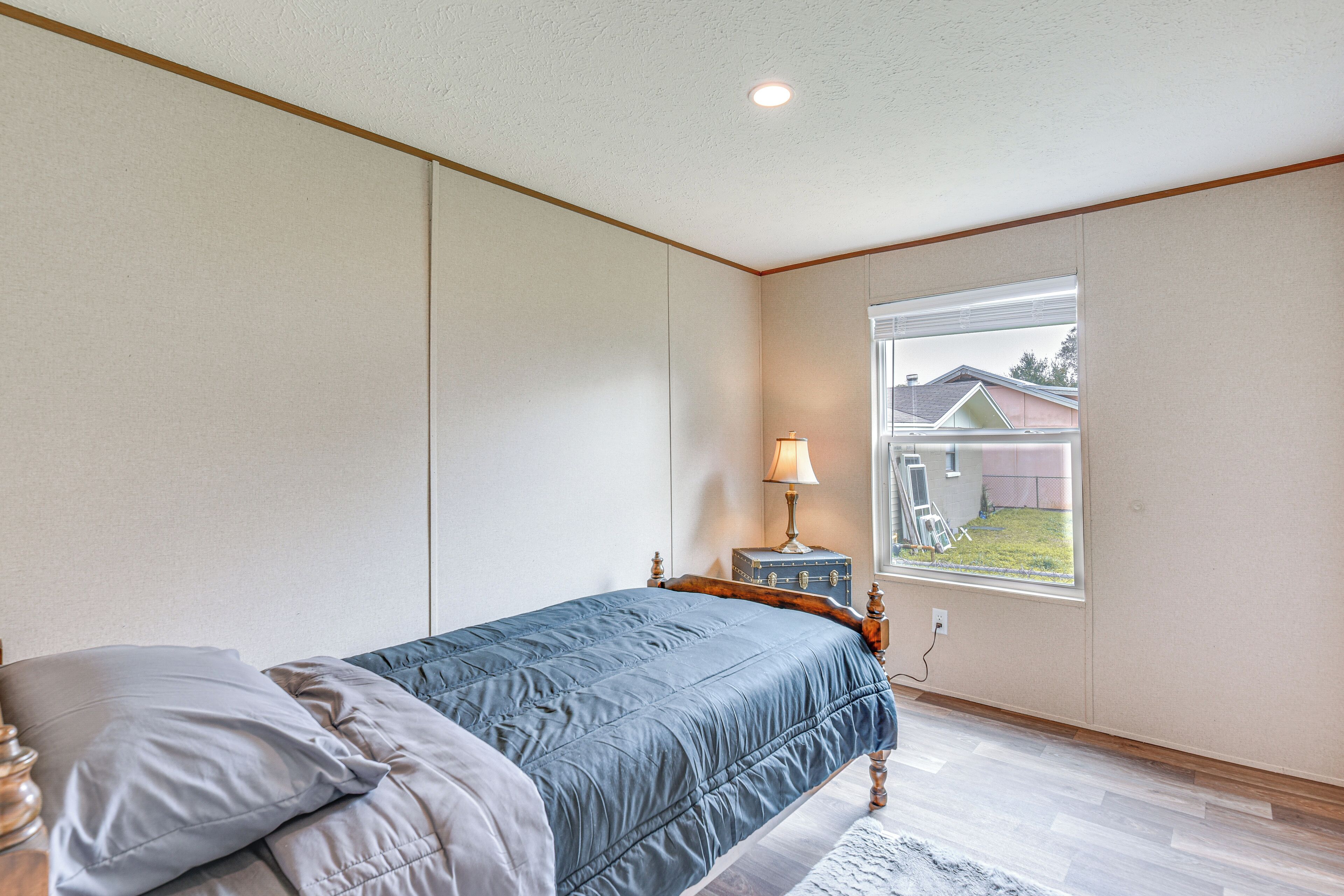 Un dormitorio con cama, mesita de noche, lámpara y ventana con vistas a una casa.