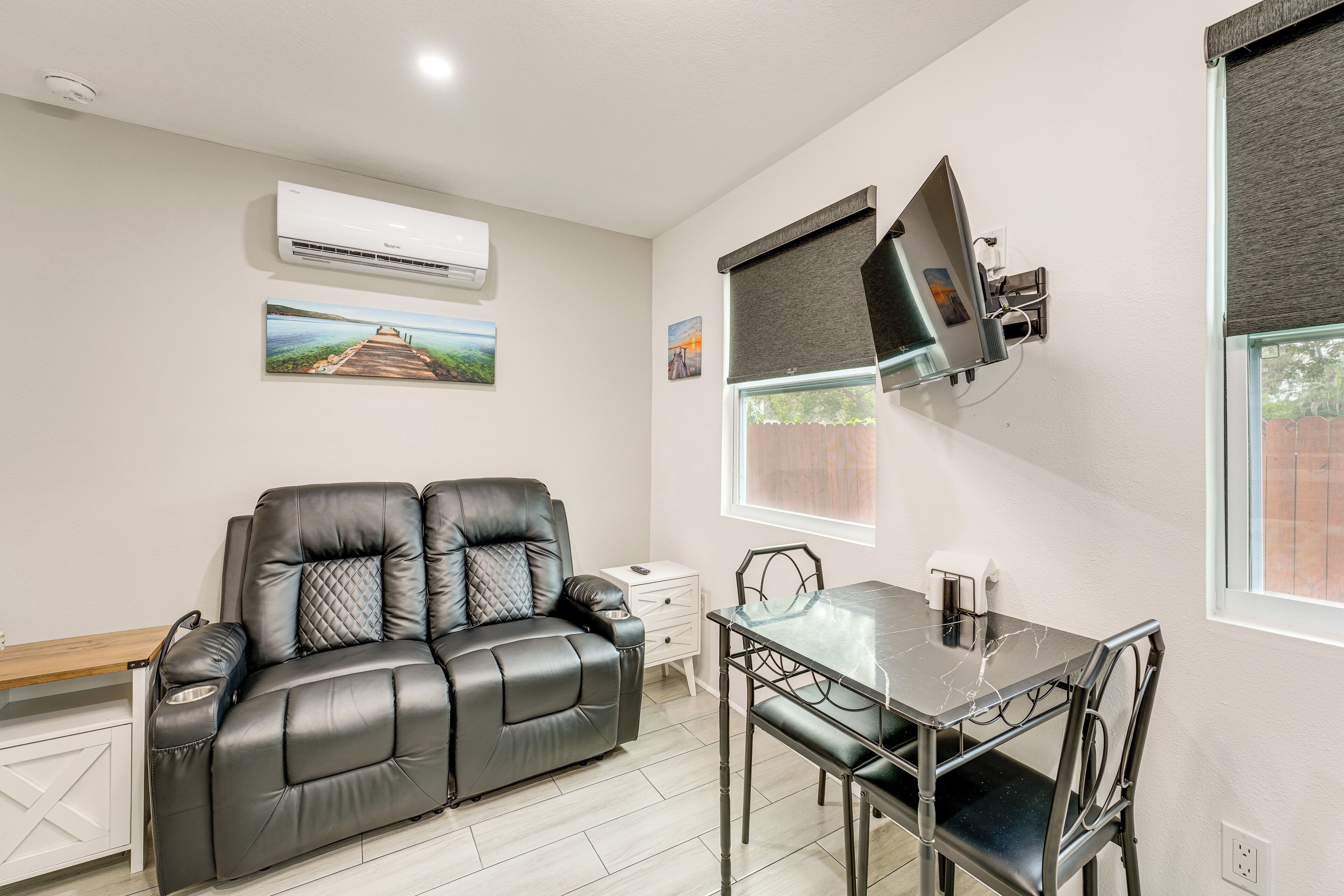 A modern living room with a leather sofa, a dining table with chairs, a flat-screen TV mounted on the wall, and a small side table.