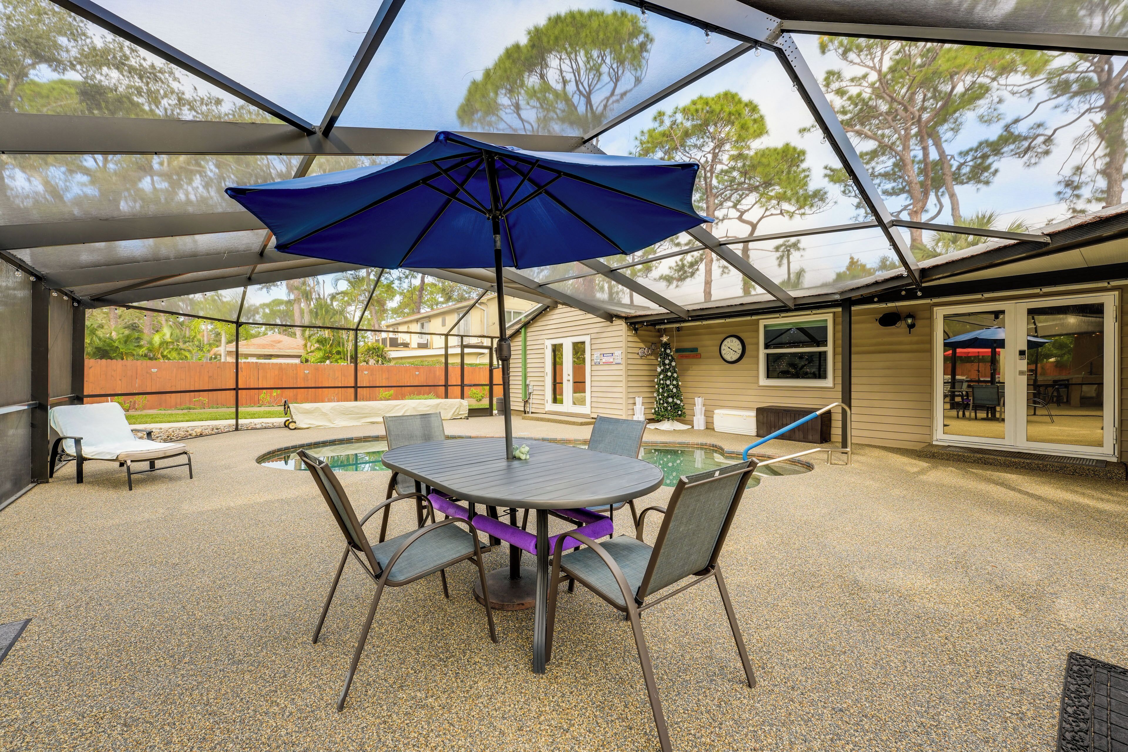 An outdoor patio with a table and chairs, a blue umbrella, and a pool in the background.