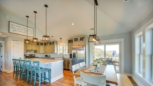 A modern kitchen with a central island, bar stools, a dining table with chairs, pendant lighting, and a view through large windows. - Luxe Family Home w/ Seasonal Pool in Duck! (Kitty Hawk)