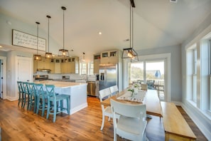 A modern kitchen with a central island, bar stools, a dining table with chairs, pendant lighting, and a view through large windows.
