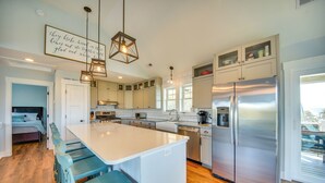 A modern kitchen with a central island, stainless steel appliances, and a dining area with blue chairs. - Luxe Family Home w/ Seasonal Pool in Duck! (Kitty Hawk)