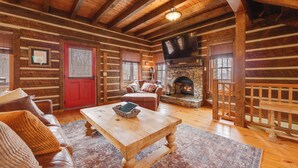 A log cabin interior with a stone fireplace, wooden furniture, and a television mounted on the wall. - Luxury Mountain Retreat w/ Hot Tub in Franklin! (Franklin)