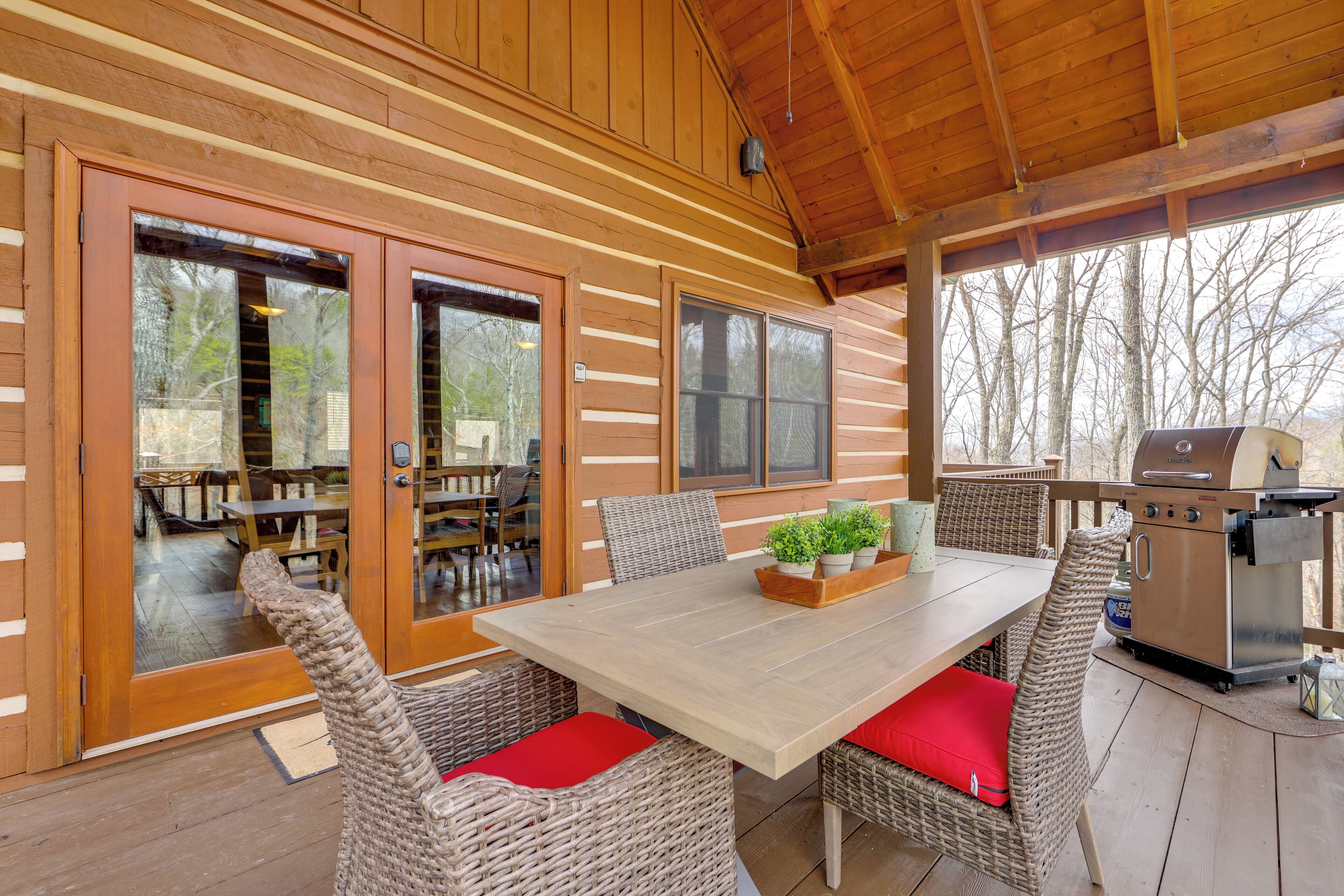 A covered outdoor dining area with wicker chairs and a table, a grill, and a view of the surrounding trees.