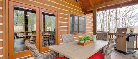 A covered outdoor dining area with wicker chairs and a table, a grill, and a view of the surrounding trees.