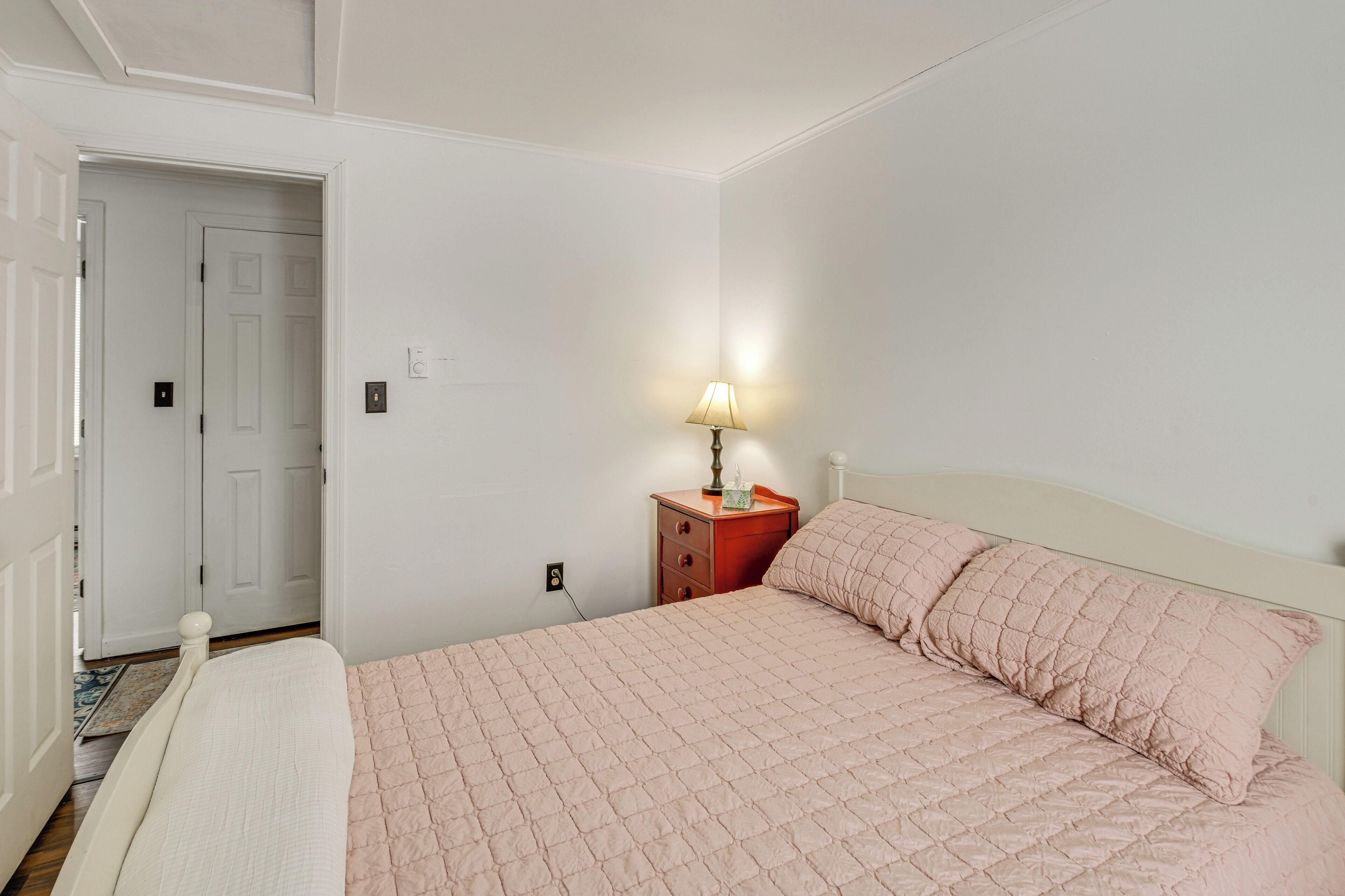 A bedroom with a bed, a nightstand with a lamp, and a door leading to another room.