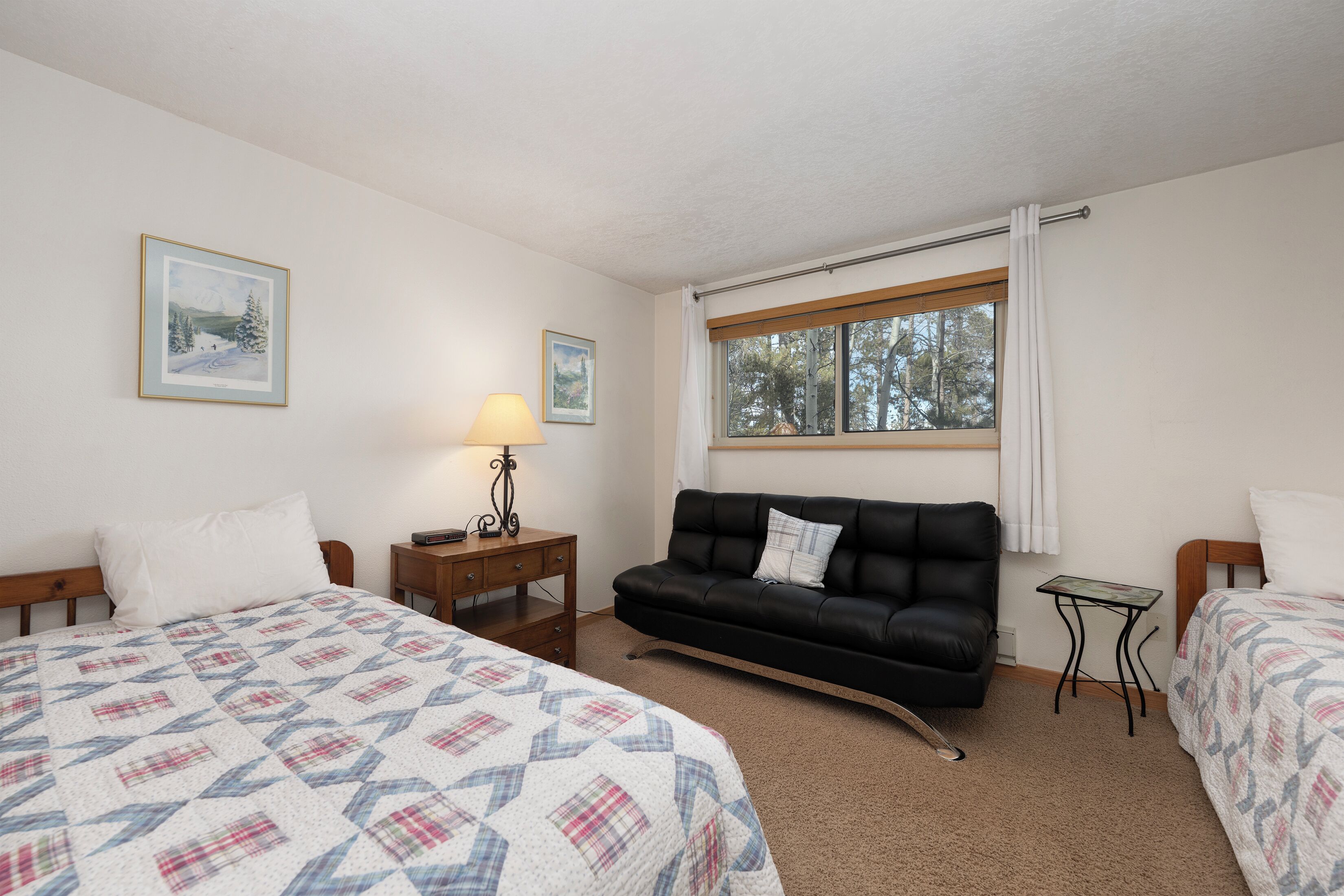 A bedroom with two beds, a black leather sofa, a small table with a lamp, and a window with curtains.