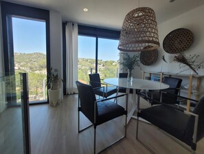 Een moderne eetruimte met een ronde tafel en zwarte stoelen, grote ramen met uitzicht op een glooiend landschap, een hangende geweven lamp en decoratieve muurkunst.
