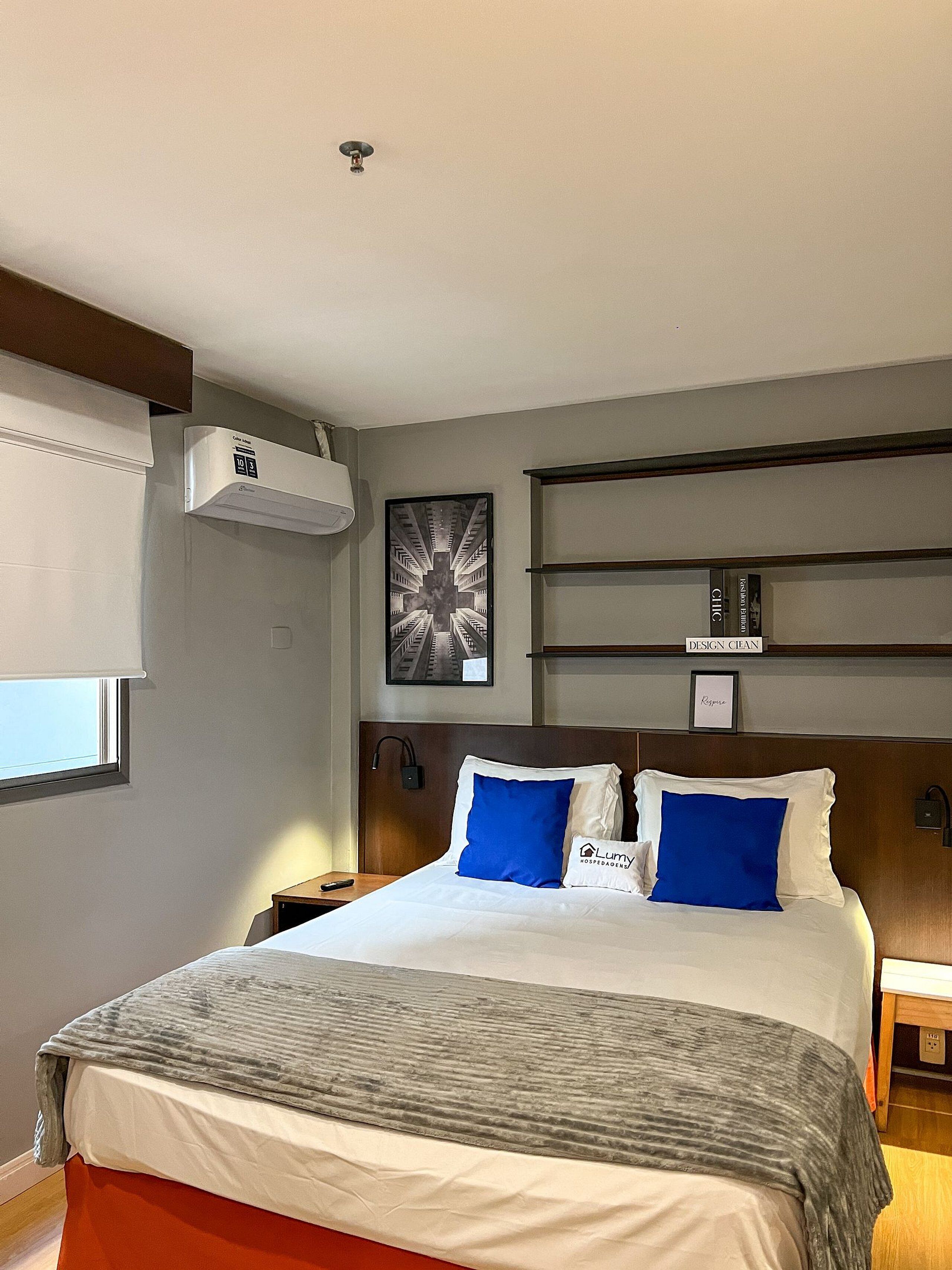 Una habitación de hotel con cama, mesita de noche, aire acondicionado de pared, un cuadro enmarco y una ventana con persianas.