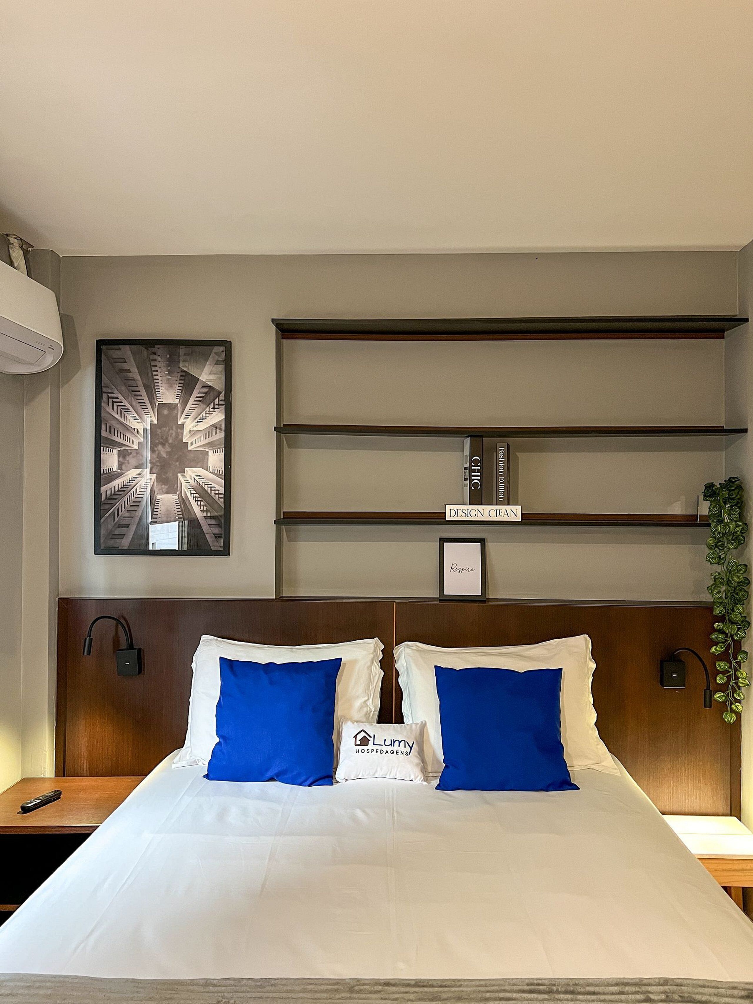 Habitación de hotel con una cama, un cuadro en la pared, un estante con un letrero y una planta.