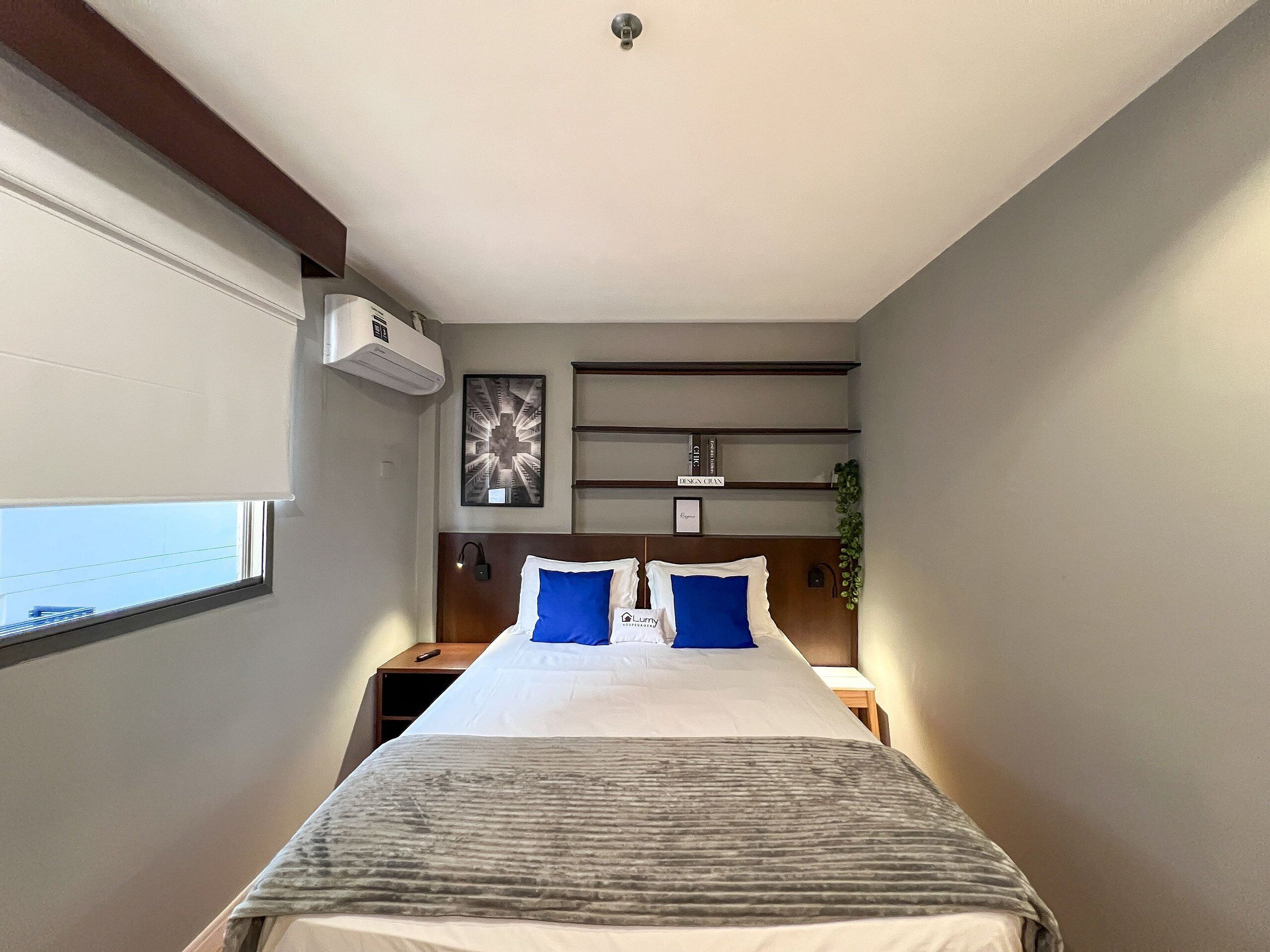 Ein Hotelzimmer mit Bett, Nachttischen, einer Klimaanlage, einem Wandregal mit Gegenständen und einem Fenster.