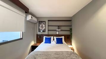 Ein Hotelzimmer mit Bett, Nachttischen, einer Klimaanlage, einem Wandregal mit Gegenständen und einem Fenster.