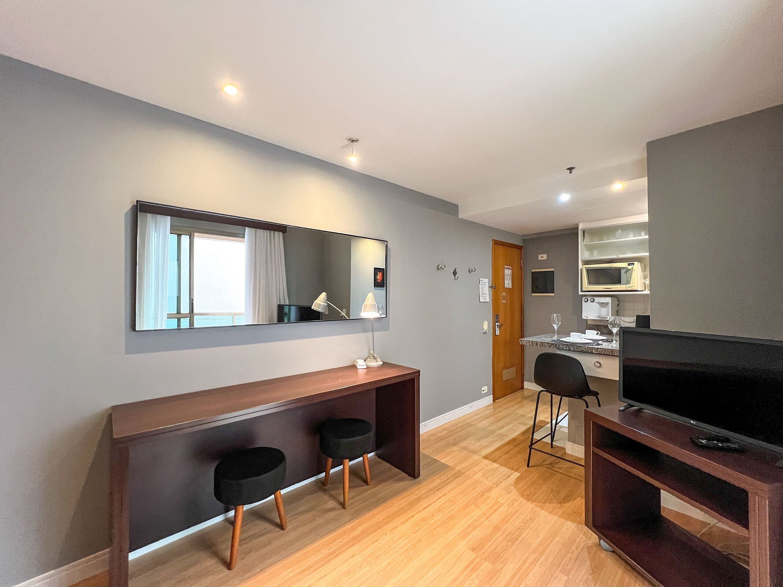 Hotelkamer met een houten bureau, twee zwarte stoelen, een grote spiegel, een televisie, een kitchenette met een magnetron en een deur.