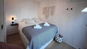 A bedroom with a bed, a bedside table with a lamp, a wall-mounted air conditioner, and a woven wall hanging.