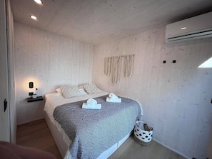A bedroom with a bed, a bedside table with a lamp, a wall-mounted air conditioner, and a woven wall hanging.