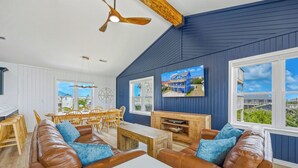 A spacious living area with a high ceiling, a dining table with chairs, and a large window offering a view of the outside. - Brand New Semi-Oceanfront Waves Home with Private Pool, Hot Tub and Game Room (Waves)