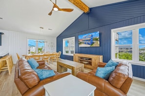 A spacious living area with a high ceiling, a dining table with chairs, and a large window offering a view of the outside. - Brand New Semi-Oceanfront Waves Home with Private Pool, Hot Tub and Game Room (Waves)